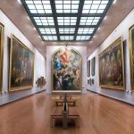 Une galerie d'art spacieuse avec un haut plafond en verre, un plancher en bois et de grandes peintures classiques dans des cadres dorés sur des murs blancs. Des bancs sont disposés au centre pour les visiteurs. Idéal pour les séjours de moyenne durée ou pour ceux qui recherchent des services de conciergerie haut de gamme. | 2BeApart Lyon