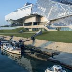 Un bâtiment moderne en verre et en métal au design anguleux se dresse au bord de l'eau à Lyon, où deux bateaux sont amarrés le long d'une digue en pierre sous un ciel bleu limpide - parfait pour les propriétaires exigeants à la recherche d'une conciergerie haut de gamme. | 2BeApart Lyon