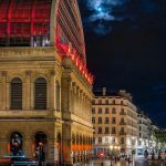 Un grand bâtiment illuminé avec des fenêtres arquées et un toit incurvé brille de lumières rouges la nuit ; les rues humides reflètent les lumières, ce qui est idéal pour les propriétaires exigeants qui recherchent des séjours de moyenne durée sous une pleine lune au-dessus des bâtiments de la ville avoisinante. | 2BeApart Lyon