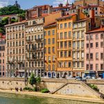Des bâtiments historiques colorés bordent une promenade fluviale sous un ciel partiellement nuageux, où des propriétaires exigeants se promènent au bord de l'eau et où la verdure recouvre la colline à l'arrière-plan. | 2BeApart Lyon