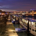 Bateaux d'excursion amarrés le long de la Seine à Paris au crépuscule, avec les lumières de la ville se reflétant sur l'eau et un ciel coloré au-dessus de la tête. Les arbres bordent la promenade et les bâtiments éloignés sont illuminés, ce qui est idéal pour les propriétaires exigeants. | 2BeApart Lyon