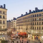 Une rue animée à la tombée de la nuit, avec des gens qui marchent, des bâtiments classiques de style européen et des magasins très éclairés, notamment des magasins de vêtements et de lunettes, qui bordent les deux côtés, capturant l'atmosphère vibrante de la location meublée de Lyon et de la gestion locative. | 2BeApart Lyon