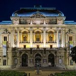 Bâtiment de théâtre illuminé la nuit, présentant une architecture ornée d'arcs, de colonnes et de balcons. Des arbres et des bannières encadrent l'entrée, où des escaliers mènent à de grandes portes en bois sous un toit en dôme - idéal pour une gestion locative ou une conciergerie haut de gamme. | 2BeApart Lyon