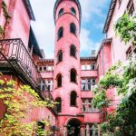 Une haute tour cylindrique rose aux fenêtres cintrées se dresse au milieu d'une cour entourée de bâtiments roses, de plantes vertes et de balcons - un cadre élégant idéal pour des expériences de conciergerie haut de gamme de moyenne durée. | 2BeApart Lyon