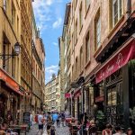 Une rue pavée animée de Lyon, bordée de grands immeubles colorés, de cafés en plein air et de gens qui profitent de la journée ; un enfant avec des ballons se tient au centre sous un ciel bleu. Parfait pour les propriétaires exigeants recherchant une conciergerie haut de gamme. | 2BeApart Lyon